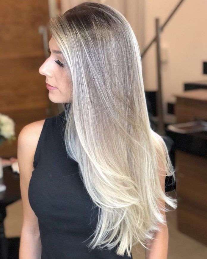 Loiro Tendência 2024: Mulher com cabelo loiro longo e liso apresentando raízes escuras e pontas platinadas, vestindo um elegante vestido preto, perfilada em um ambiente interno sofisticado.