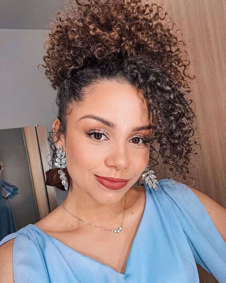 Mulher com cabelo cacheado estilizado em um coque abacaxi, um penteado alto e elegante, perfeito para destacar cachos naturais.