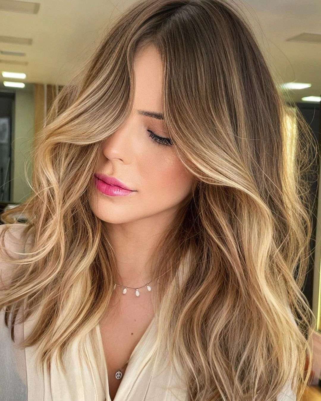 Loiro Tendência 2024: mulher com cabelo ondulado em tons de loiro mel que se destacam, complementando sua maquiagem suave e batom rosa.