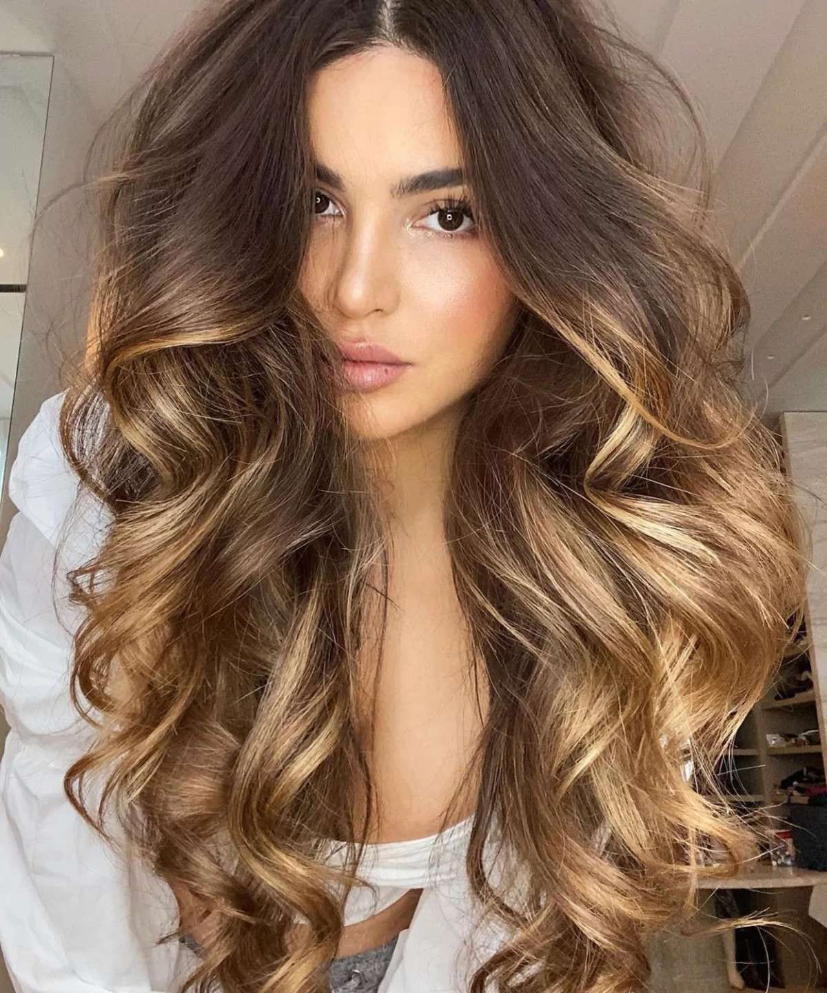 Loiro Tendência 2024: jovem com cabelo castanho ao loiro ombré, ondas volumosas, vestindo camisa branca, exibindo beleza natural e maquiagem suave.